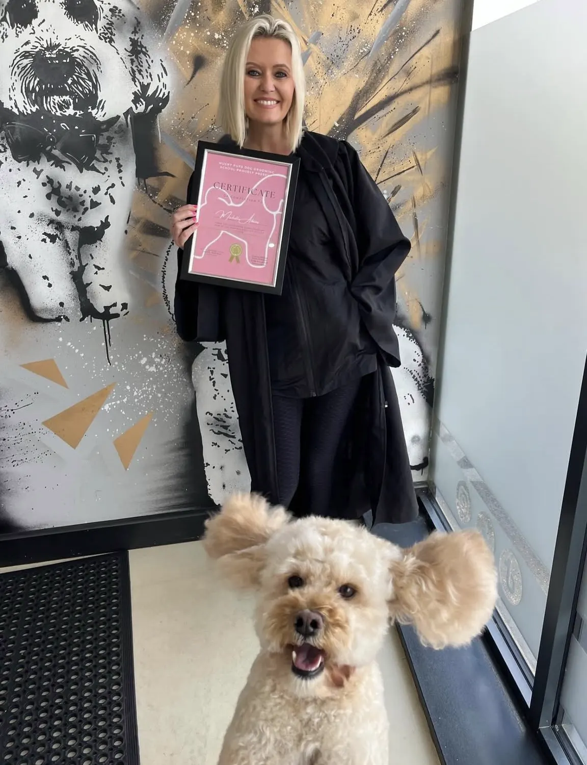 Michelle Adams holding her grooming qualification certificate with a happy cream poodle-cross in front